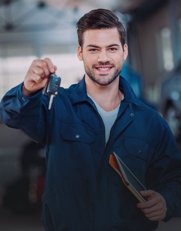 Smiling male mechanic in blue coveralls holds up car keys toward the camera, clipboard tucked under his arm in a workshop.