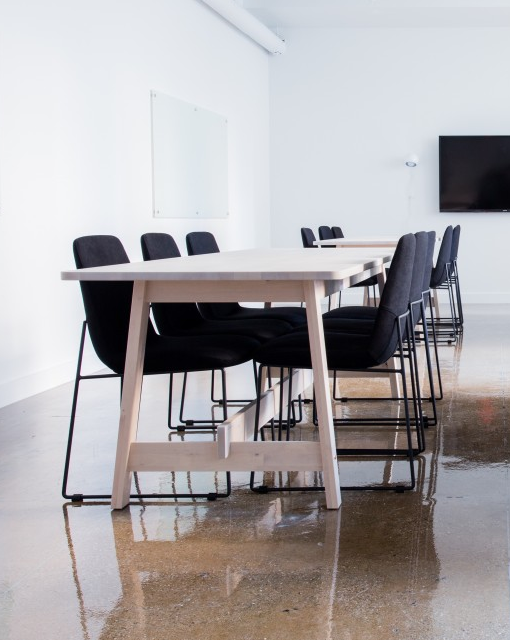 Conference room with a long light-wood table and black chairs against white walls, a whiteboard, and a mounted screen in the back.